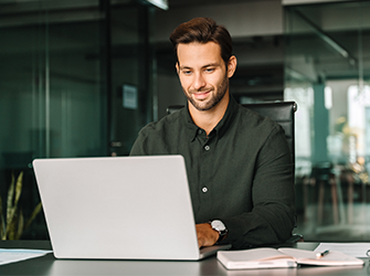 A man with dark brown hair and a beard on a laptop at a desk.