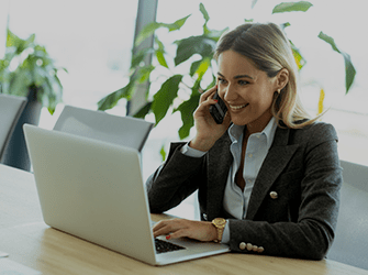 A professionally dressed woman sat at a desk smiling whilst talking on the phone.