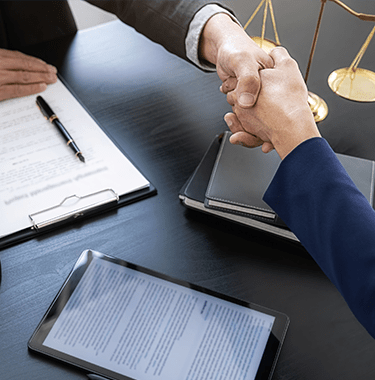 A close up of two men in suits shaking hands across a desk with documents and and ipad on there.