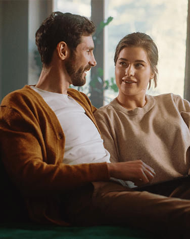 A couple sat on a couch together looking at one another whilst talking.