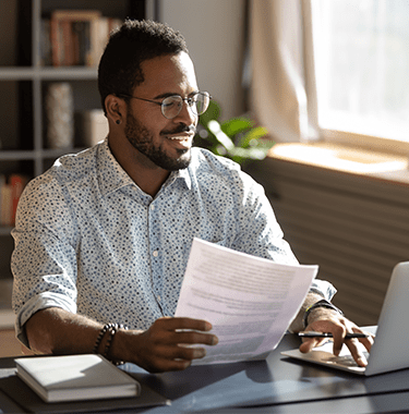A man sat at a desk looking at a laptop with documents in his hand.