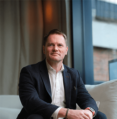 A mid-shot of Scott Clay sat on a white couch looking into the distance.