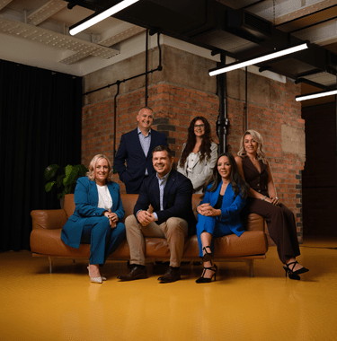 A wide shot of the senior leadership team sitting on a leather couch in front of a red brick wall.