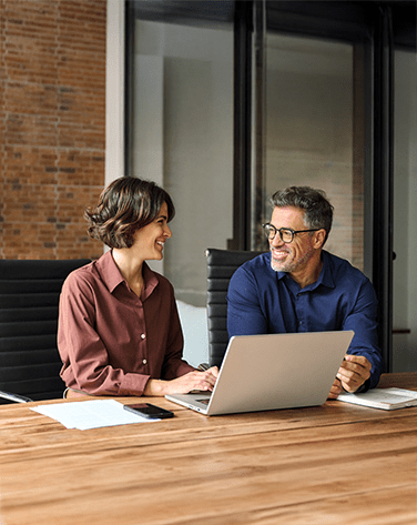 A woman and a man sat behind a desk with a laptop on it talking.