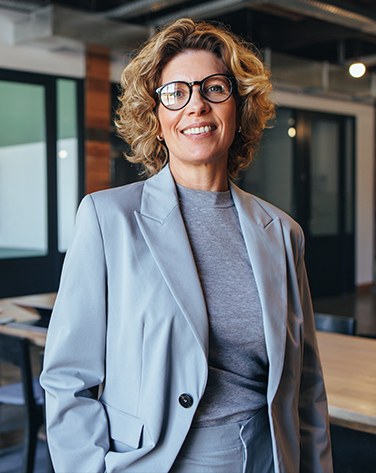 A woman in a grey blazer and jumper smiling with glasses and short blonde hair.