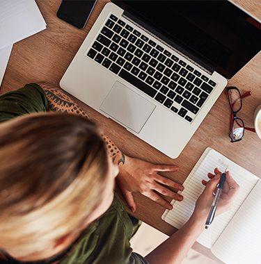 A woman writing on a notepad at a desk with a laptop in front of her.