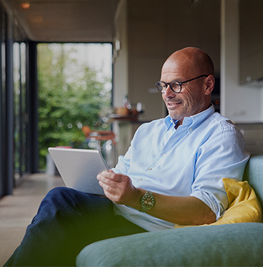 An older bald man sat on a couch on an iPad in a luxury house.
