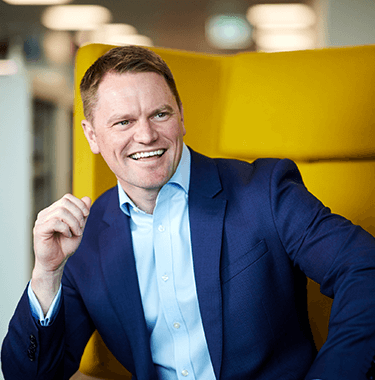 A man with short brown hair smiling wearing a navy blue suit sitting on a yellow chair v2