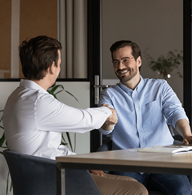Two professional men sat at a table laughing whilst shaking hands.