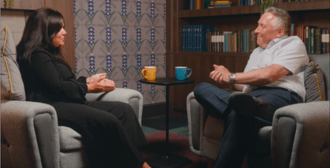 A man and woman sat on comfortable chairs talking in front of a bookcase and fireplace.
