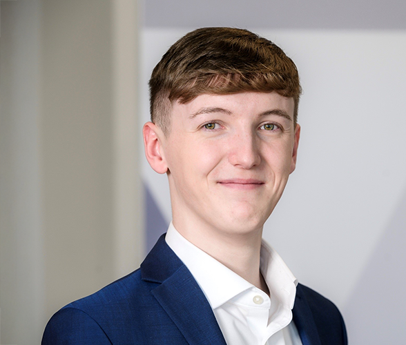 A young man smiling with short brown hair wearing a navy blue blazer and white shirt.