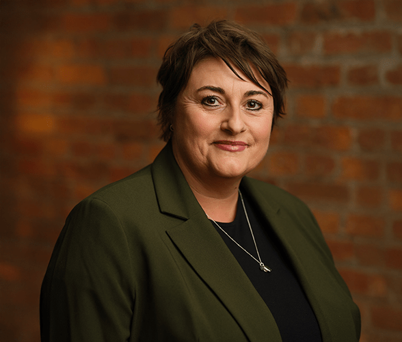 Jo Elton with short brown hair smiling in front of a red brick wall.